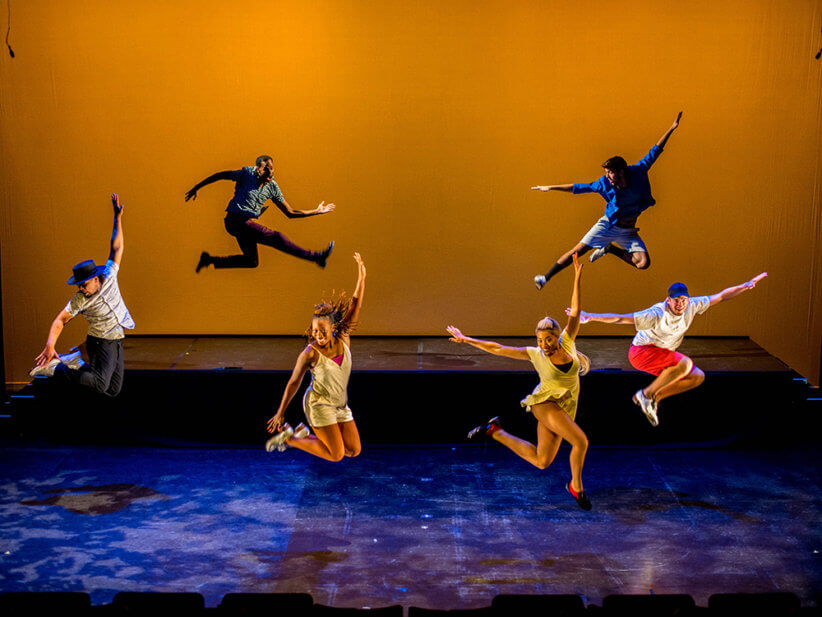 a group of six dancers holding their arms out mid-jump on stage