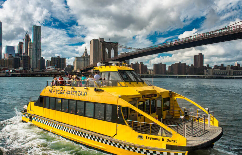 a yellow New York Water Taxi sits in the water with the Brooklyn Bridge and city skyline in the background