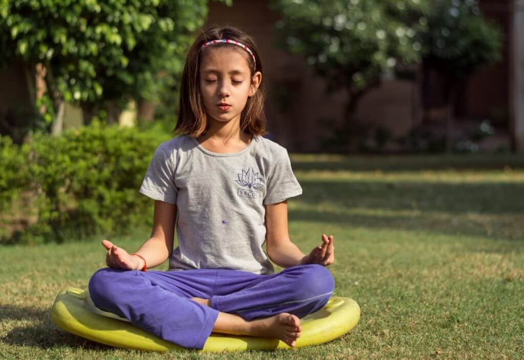 a girl sits cross-legged on a yellow mat on the grass with her eyes closed as she meditates
