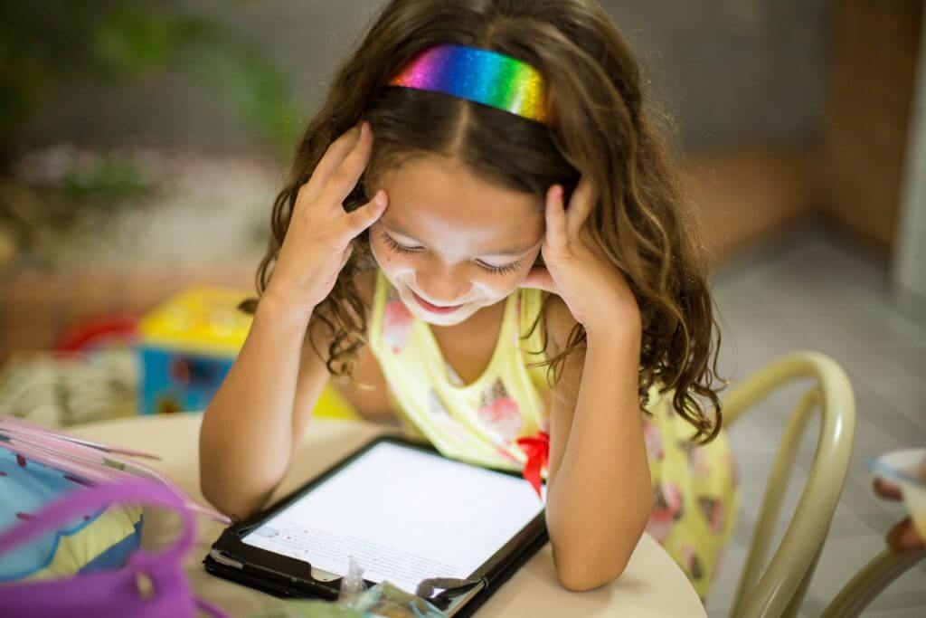 a girl with a rainbow headband smiles as she looks down at the screen of a tablet, free tutoring