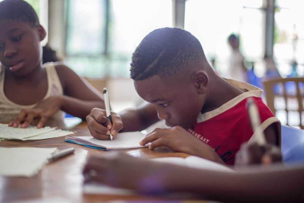 a boy writes on a paper at a table next to another child 