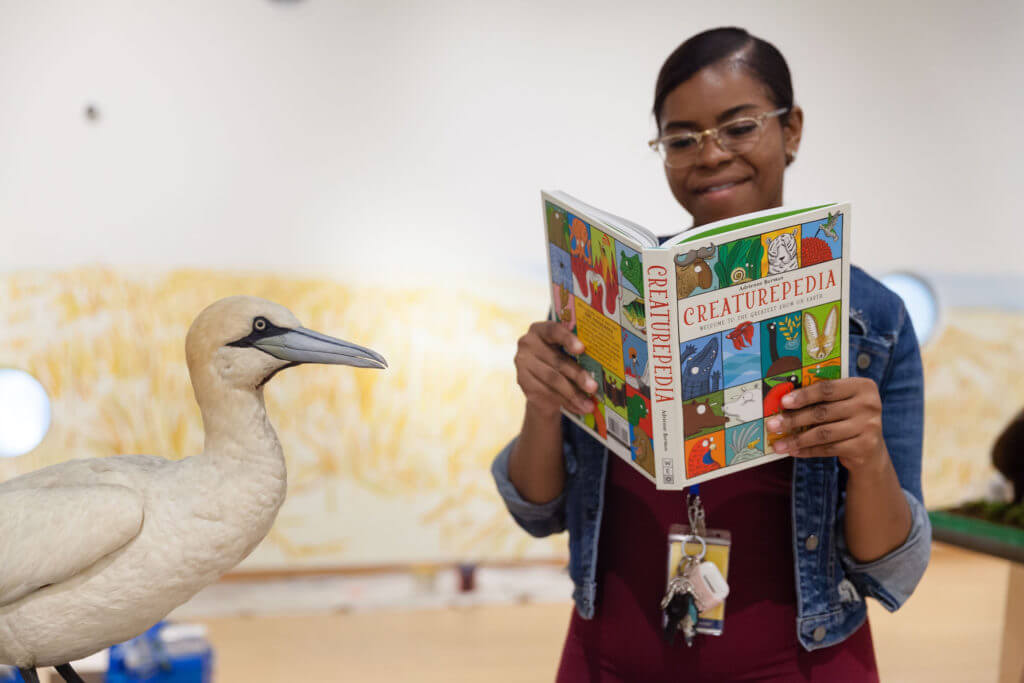 a woman next to a stuffed white bird reads a book called "Creaturepedia"