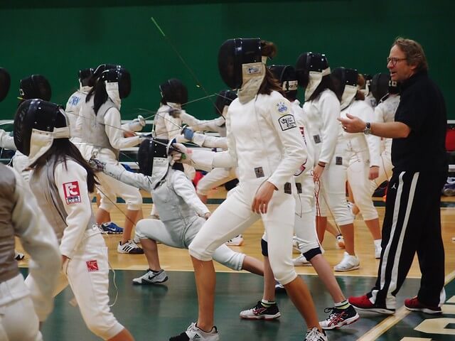 people in white clothing fencing as a coach in black clothing stands behind them and gives advice