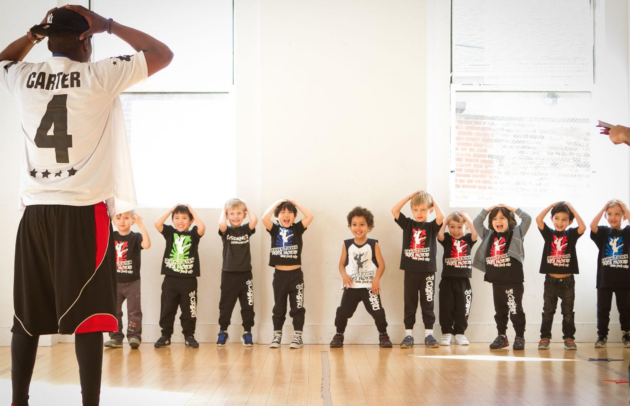 a group of boys watch and mimic a dance instructor as he puts his hands behind his head 