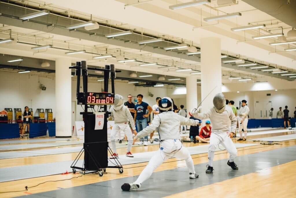 two opponents in white clothing fence each other near a digital game timekeeper while people watch on