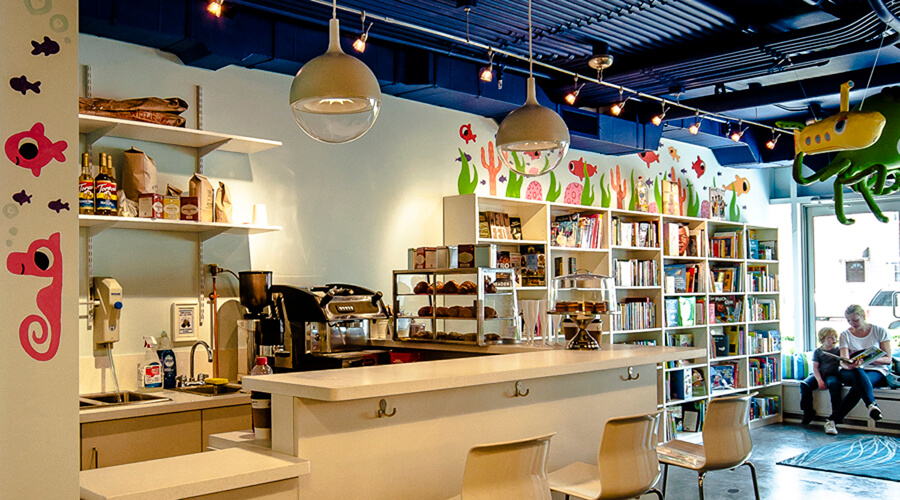 The image titled "openingpage3-2.jpg" shows a brightly lit modern cafe with a white countertop, shelves stocked with books and treats, playful wall art, and people reading by the window.