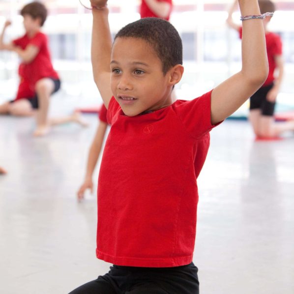 a boy in a red shirt sits on his knees with his arms in the air as other children hold a similar pose in the background