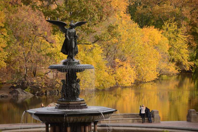 Fountain with angel statue in Central Park, surrounded by autumn trees and two people standing by a lake in the background.