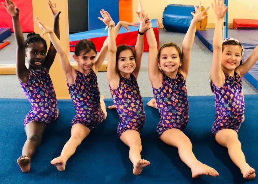Five young girls sit in a split position on a blue gymnastics mat, wearing matching floral leotards and raising their arms overhead.