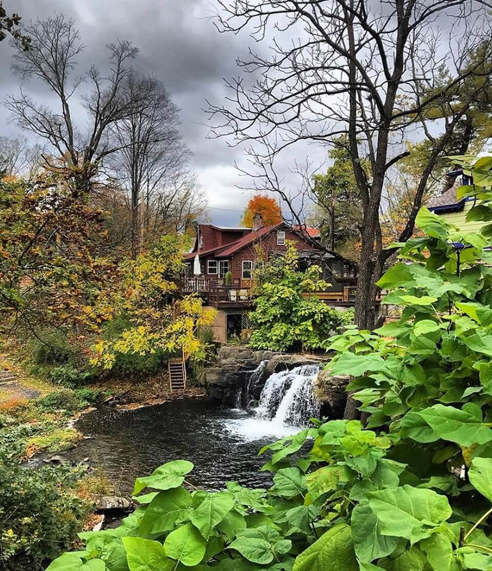 A red house sits by a small waterfall and pond, surrounded by autumn trees and greenery under a cloudy sky, creating a tranquil scene.