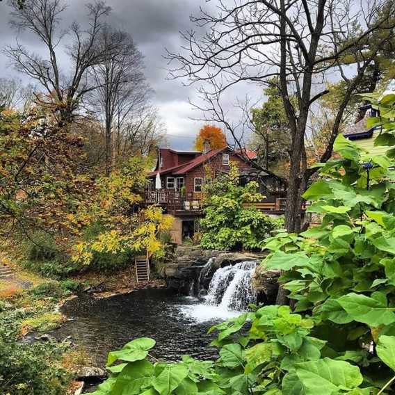 A red house sits by a small waterfall and pond, surrounded by autumn trees and greenery under a cloudy sky, creating a tranquil scene.