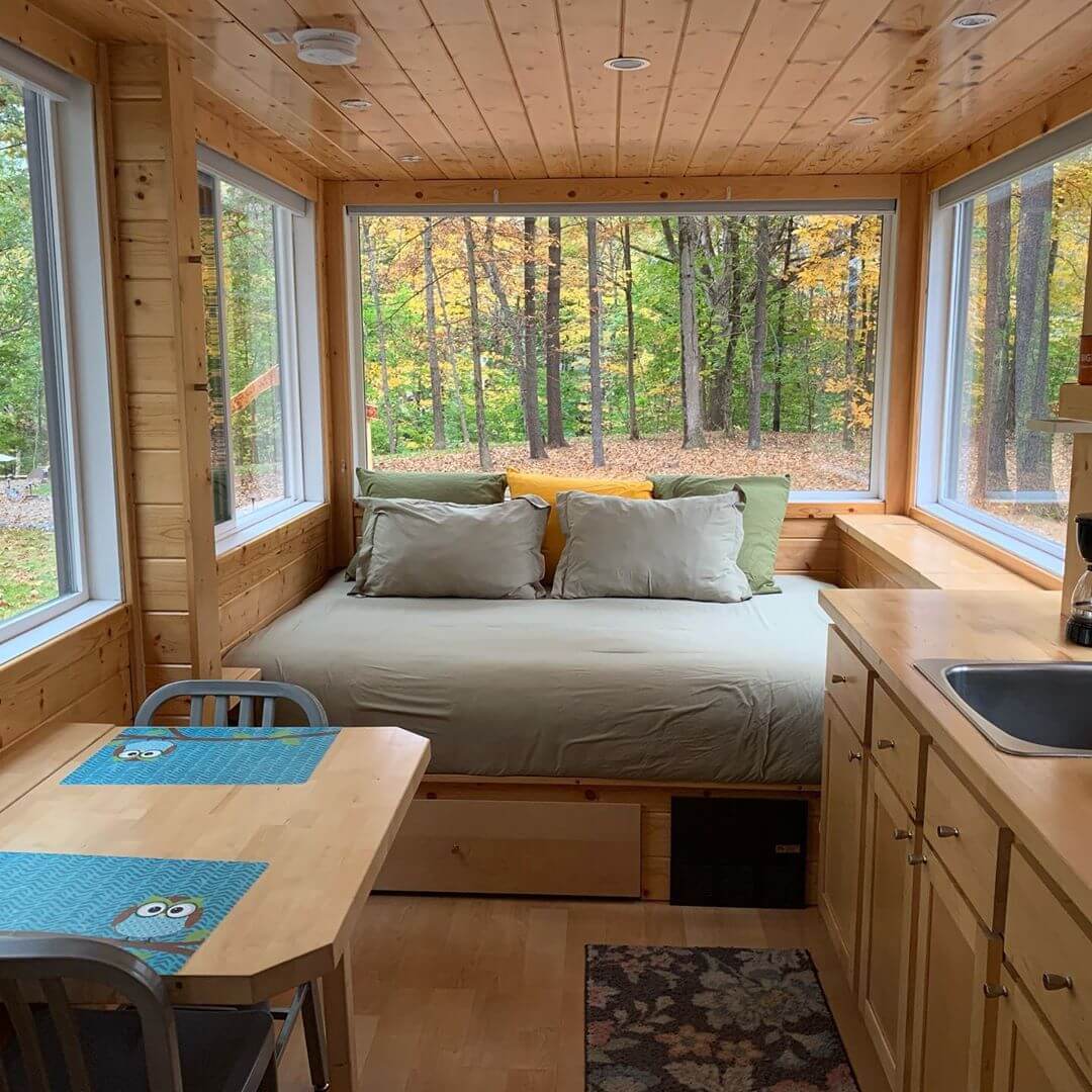 A cozy tiny home features large windows, a bed facing the forest, a compact kitchen, and a small table set with blue placemats.