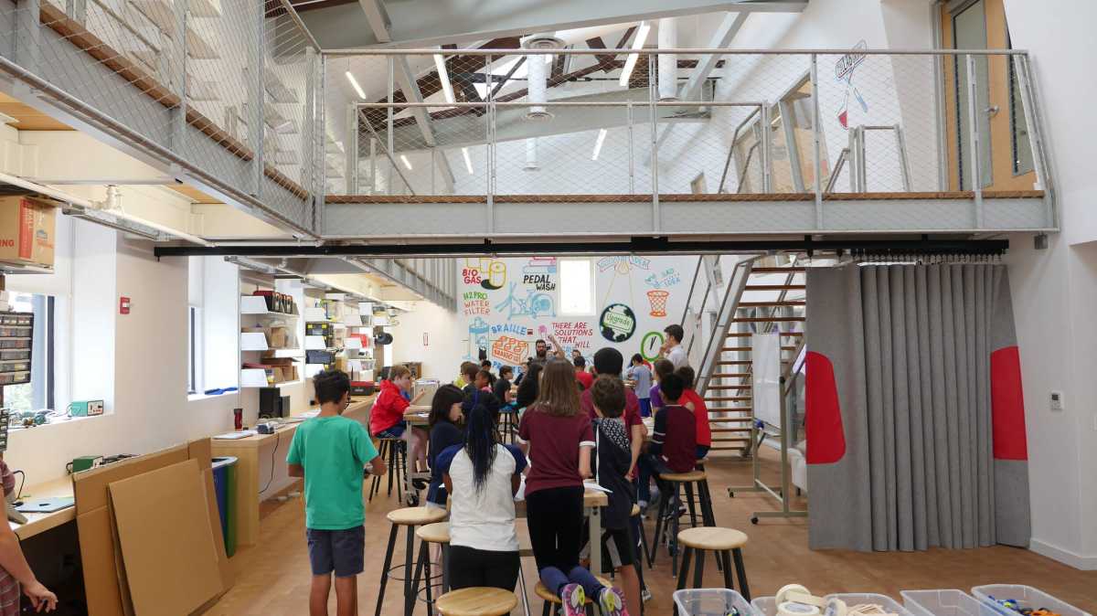 Students gather in a spacious maker lab with stools, creative workstations, and colorful murals on the walls, under a modern mezzanine.