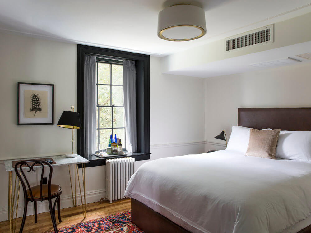 Bright bedroom with a white bed, window, desk, chair, lamp, and rug, decorated in a modern, minimalist style with soft lighting.