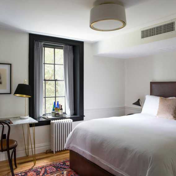 Bright bedroom with a white bed, window, desk, chair, lamp, and rug, decorated in a modern, minimalist style with soft lighting.
