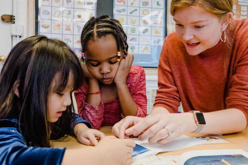 A teacher and two young students engaged in a learning activity at a table, with educational posters visible on the classroom wall behind them.