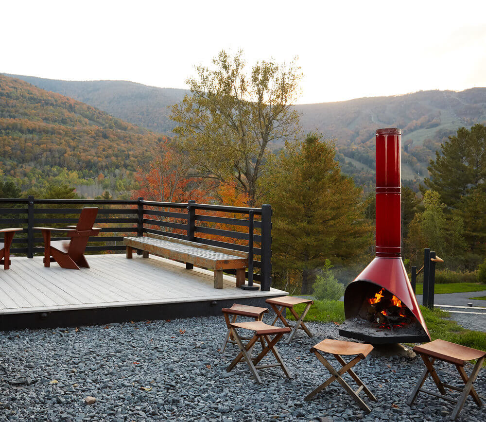 
Outdoor deck with benches and chairs overlooks scenic autumn mountains, featuring a red standing fire pit with stools on gravel. A person loads a Christmas tree onto an orange vehicle, with others standing nearby outdoors among rows of evergreen trees.