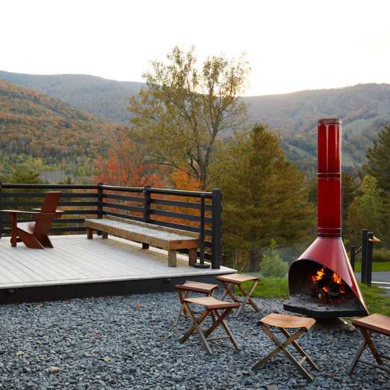 Outdoor deck with benches and chairs overlooks scenic autumn mountains, featuring a red standing fire pit with stools on gravel. A person loads a Christmas tree onto an orange vehicle, with others standing nearby outdoors among rows of evergreen trees.