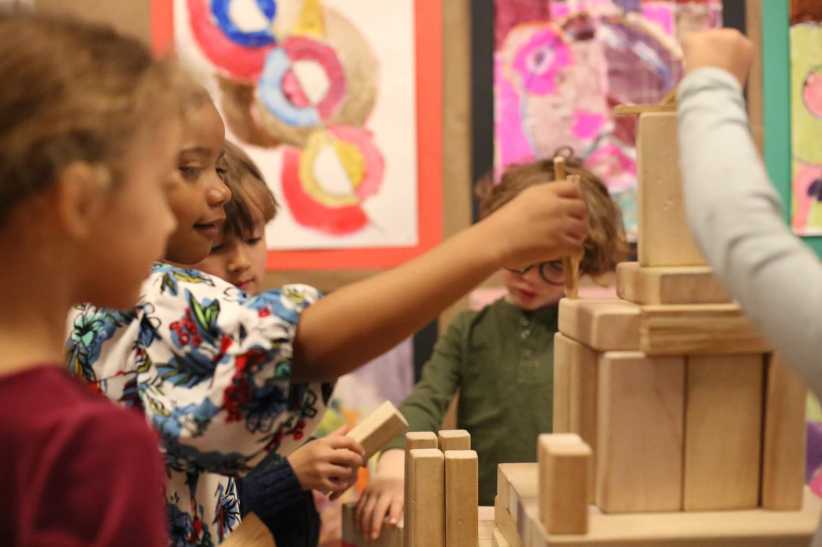 
Children building with wooden blocks in a classroom, colorful abstract artwork hangs on the walls in the background.