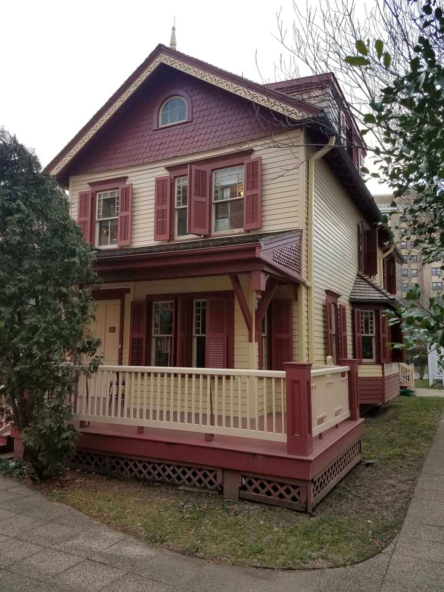 Two-story Victorian house with cream siding and maroon trim, shutters, porch, and decorative eaves, surrounded by trees and grass.