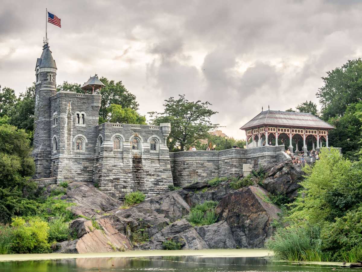 A historic stone castle with an American flag, beside a pavilion, sits on rocky terrain surrounded by trees and a pond.