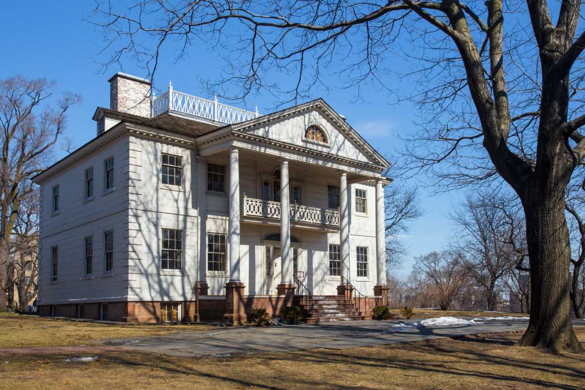 A historic white mansion with columns and balcony, surrounded by leafless trees and a lawn on a clear, sunny day.