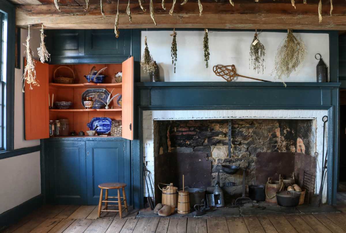 Rustic colonial kitchen with wood floors, blue-green cabinets, orange shelves, an open hearth, hanging dried herbs, and pottery.