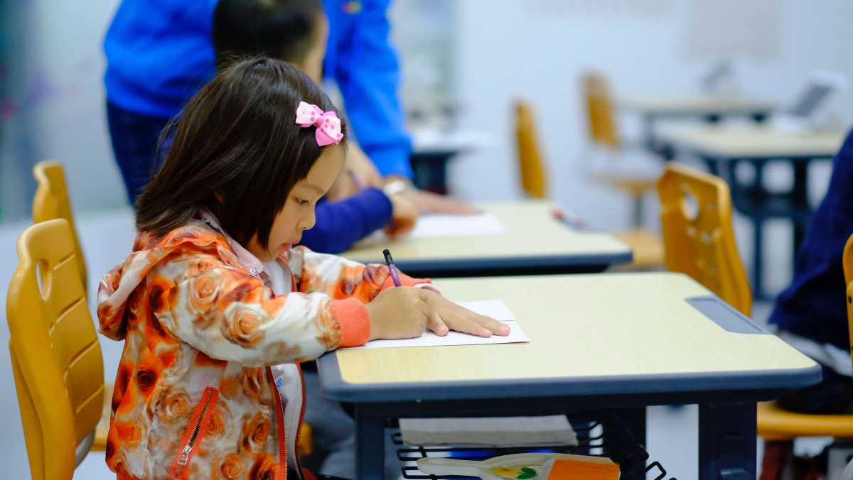 A young child wearing a floral jacket and pink hair bow writes on paper at a classroom desk, with other students in the background.
