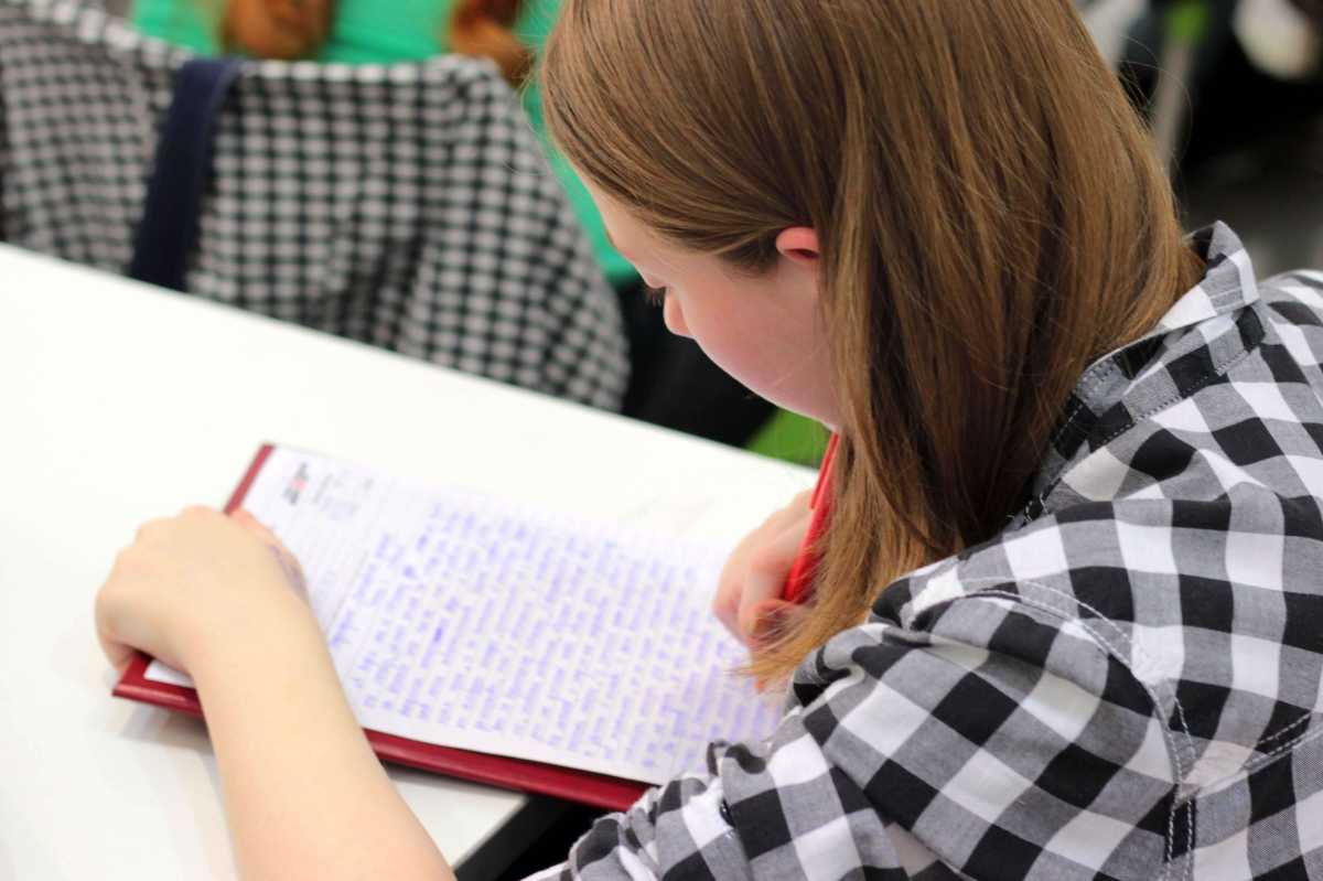 A person with long hair wearing a checkered shirt writes with a pen on a sheet of paper at a desk; face is blurred.