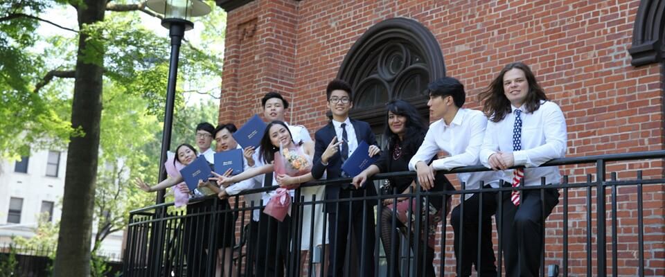 A group of students holding diplomas stand by a brick building, celebrating; their faces are blurred and one holds a bouquet of flowers.