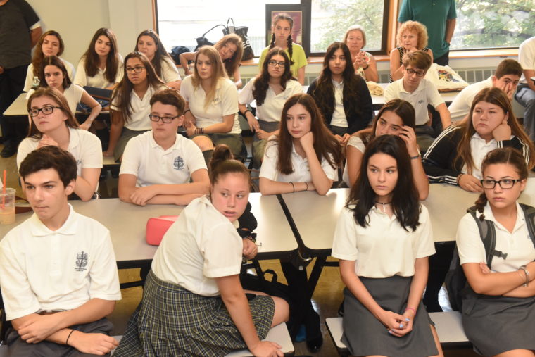A group of students in uniforms sit and stand around classroom tables, with their faces blurred, listening and talking together.