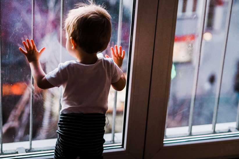 Rear view of a toddler in a white shirt and striped pants looking out a window with their hands on the glass.