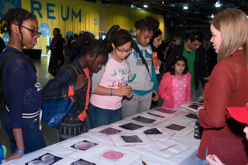A group of young girls look at scientific diagrams on a table while an instructor explains the exhibit.