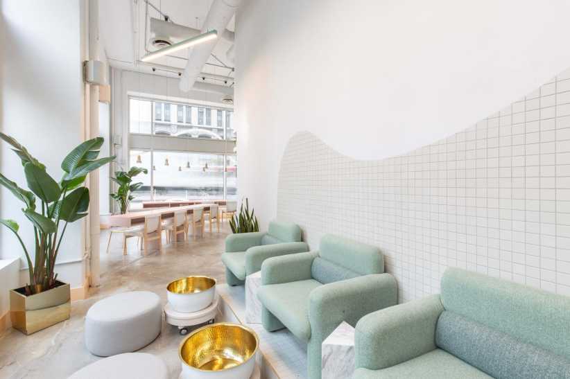 Modern minimalist lounge interior features white wavy tiled walls, large windows, lush green plants, teal modular sofas, gold pots, poufs, and a long wooden table.