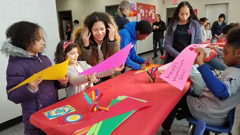 Children and adults create colorful paper crafts at a red table during a festive "Happy Valentine's Day" event.