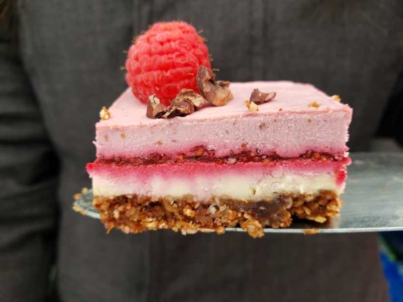 A slice of layered raspberry cheesecake with a nut crust, topped with a fresh raspberry and cacao nibs on a metal server.