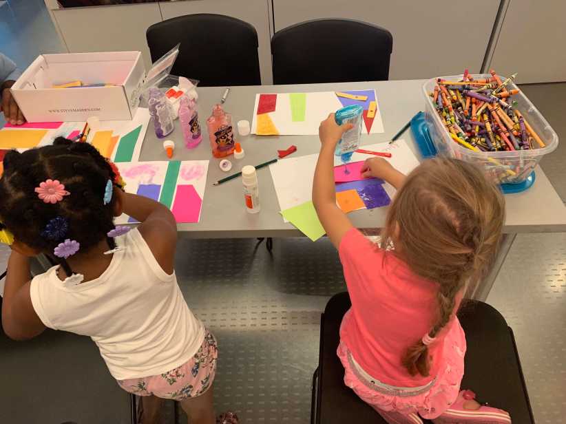 Two children at a table using glue and colorful paper to create collages near a large bin of crayons.