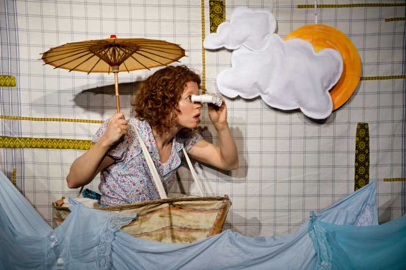 A woman in a prop boat looks through binoculars under a paper sun and clouds on a fabric backdrop.