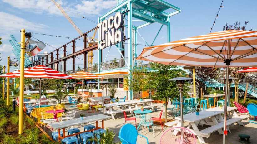 Vibrant Tacocina taco stand patio in Brooklyn features "TACOCINA" sign on turquoise structure, colorful striped umbrellas, picnic tables, chairs, plants, and construction crane under blue sky.