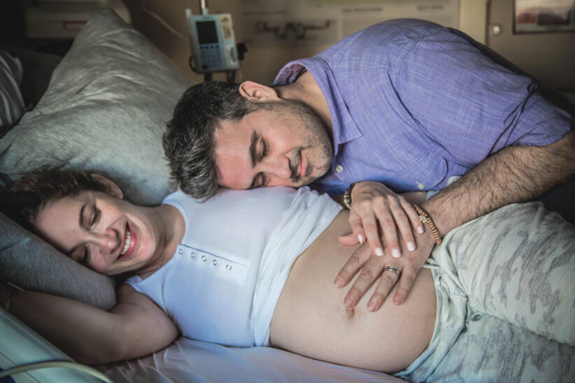 pregnant woman laying in hospital bed with husband hugging her