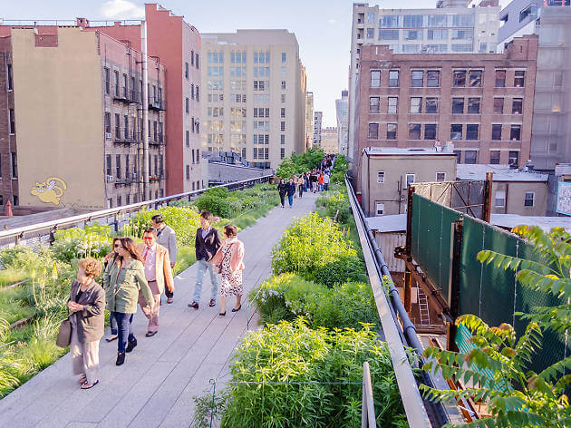 People stroll along the High Line park a raised walkway with lush plants, surrounded by brick buildings in sunny New York City.