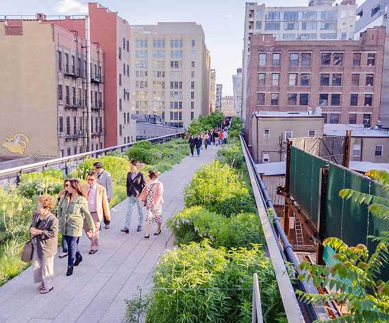 People stroll along the High Line park a raised walkway with lush plants, surrounded by brick buildings in sunny New York City.