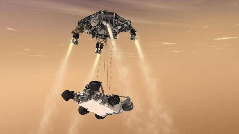 A NASA Mars rover is suspended below a sky crane as it descends toward the Martian surface, with dust clouds swirling below.