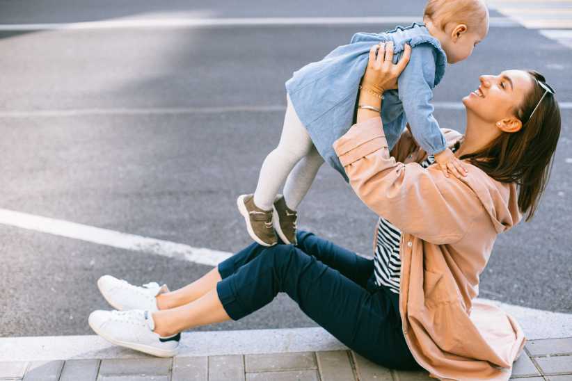 A woman sits on a curb, smiling as she lifts a baby into the air at a crosswalk, sharing a joyful moment on a quiet city street.