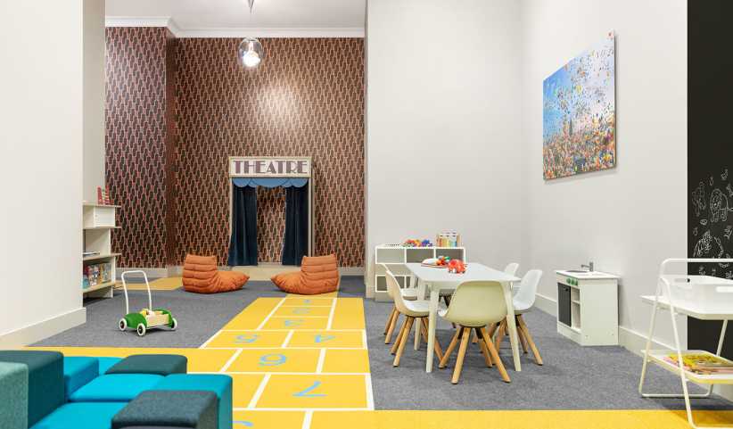 Children’s playroom with hopscotch carpet, a mini theater, small tables and chairs, bean bags, toys, and colorful artwork.