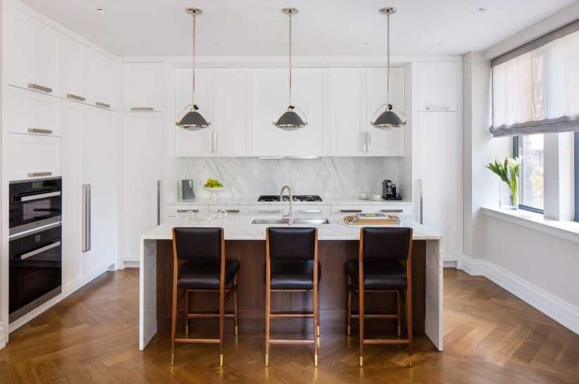 Modern kitchen with white cabinets, marble backsplash, three pendant lights, brown island, and barstools next to a sunny window.