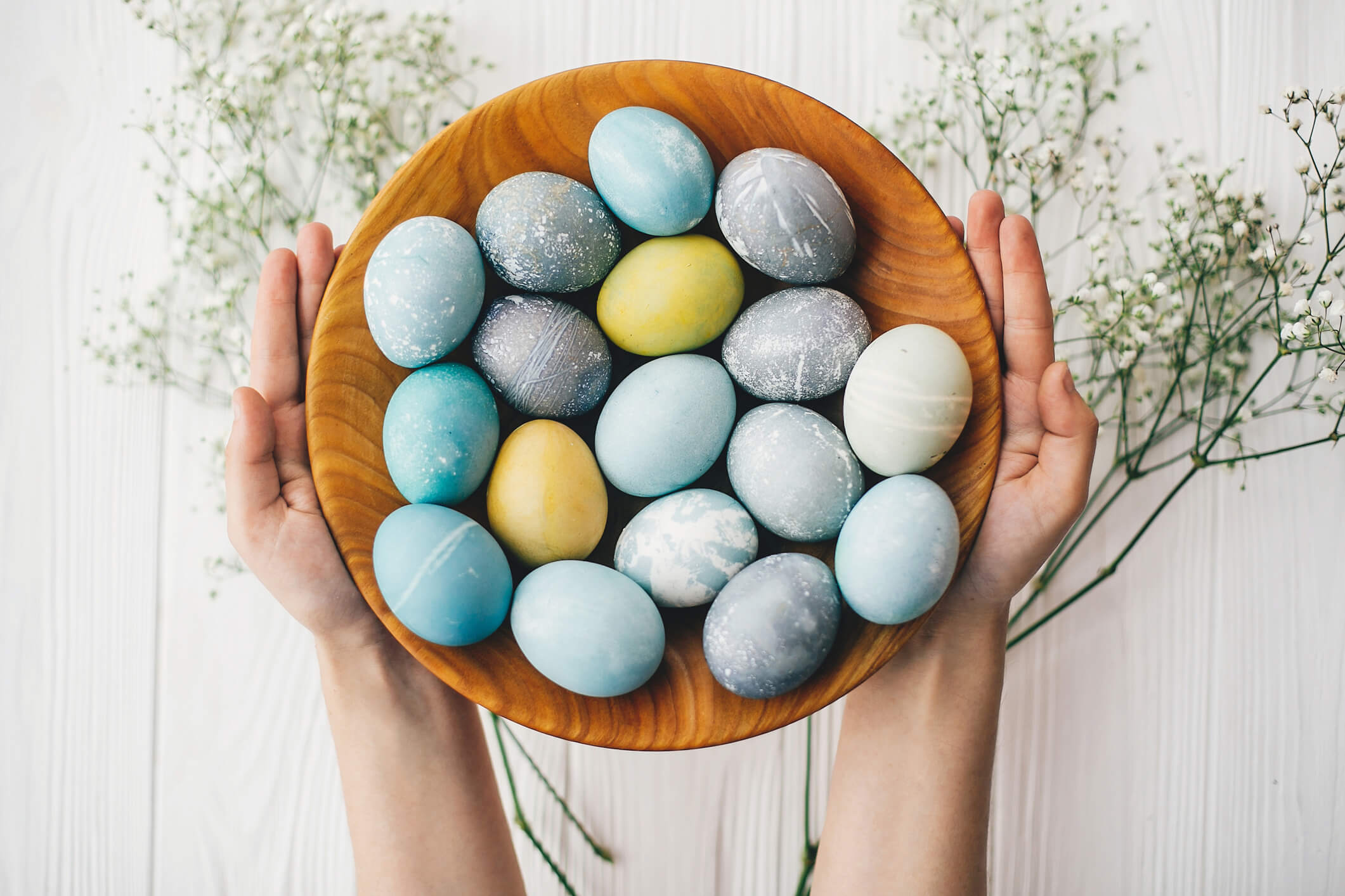 Hands holding stylish easter eggs in wooden plate with spring flowers on white wooden background. Modern easter eggs painted with natural dye in blue, grey, yellow color. Happy Easter