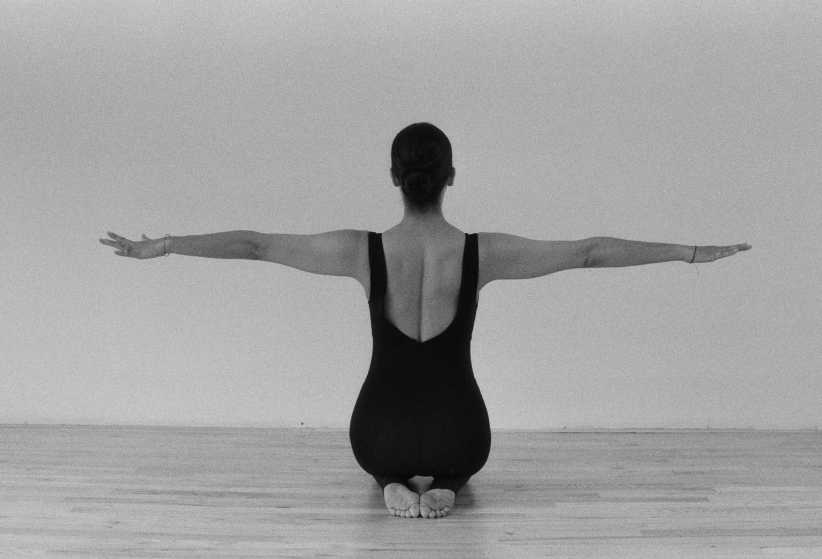 A black-and-white photo from behind of a woman in a dark bodysuit kneeling with her arms extended straight out to her sides.