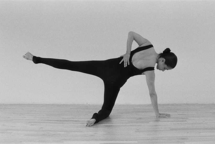 A black-and-white photo of a woman in a bodysuit balancing on one knee and hand with one leg extended.