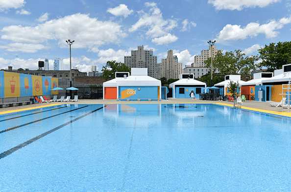 Bright blue city pool with colorful cabanas and lounge chairs under a sunny sky, framed by urban high‑rise buildings.